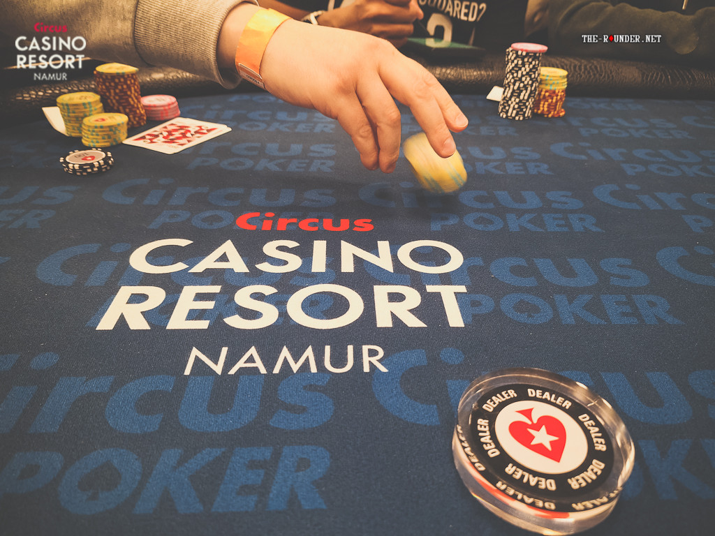 Ein professioneller Pokertisch mit grüner Filzoberfläche, umgeben von Stühlen und beleuchtet von einer Deckenleuchte. Die Spielerchips und Karten sind auf dem Tisch verteilt, während