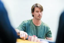 Ein Mann sitzt am Pokertisch und hält seine Karten an. Er wirkt konzentriert und trägt eine dunkle Brille. In der Mitte des Tisches liegen Pokerchips und eine Karte mit dem