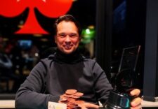 Ein lachender junger Mann, der eine schwarze Sonnenbrille trägt und vor einem verschwommenen Hintergrund posiert. Er hat kurze dunkle Haare und trägt ein graues T-Shirt.