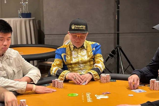 Ein Mann in dunkler Kleidung sitzt an einem Pokertisch und betrachtet konzentriert seine Karten. In seiner Hand hält er Pokerchips, während er von anderen Spielern umgeben ist.