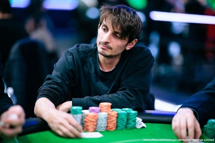 Ein Mann in einem dunklen Anzug sitzt an einem Pokertisch und betrachtet konzentriert seine Karten. Im Hintergrund sind andere Spieler und Dealer zu sehen.