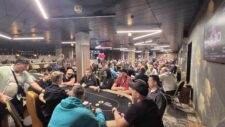 Ein Mann in einem schwarzen Anzug sitzt an einem Pokertisch und hält eine Hand mit Spielkarten. Im Hintergrund sind weitere Spieler und Chips zu sehen.