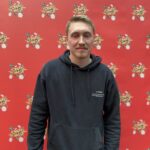 Ein Mann sitzt an einem Pokertisch und hält Karten in der Hand. Er trägt eine schwarze Kappe und konzentriert sich auf das Spiel. Im Hintergrund sind Pokerchips und Getränke zu