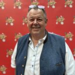 Ein Mann spielt Poker am Tisch mit anderen Spielern. Er hält Karten in der Hand und schaut konzentriert auf den Tisch. Ein Stapel Pokerchips liegt vor ihm.