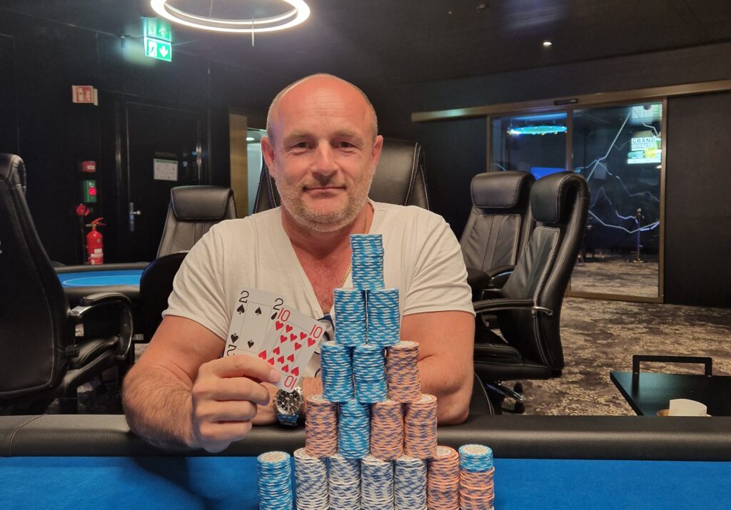 Ein professionell gestalteter Pokertisch mit grünem Filz, umgeben von Spielkarten, Pokerchips und Getränken. Ein Spieler mit Sonnenbrille und Kappe sitzt am Tisch und studiert
