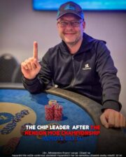 Ein Mann mit dunklen Haaren und Bart sitzt an einem Pokertisch und hält zwei Karten in der Hand. Er trägt ein schwarzes Hemd und eine schwarze Kappe. Im Hintergrund sind Pokerchips und