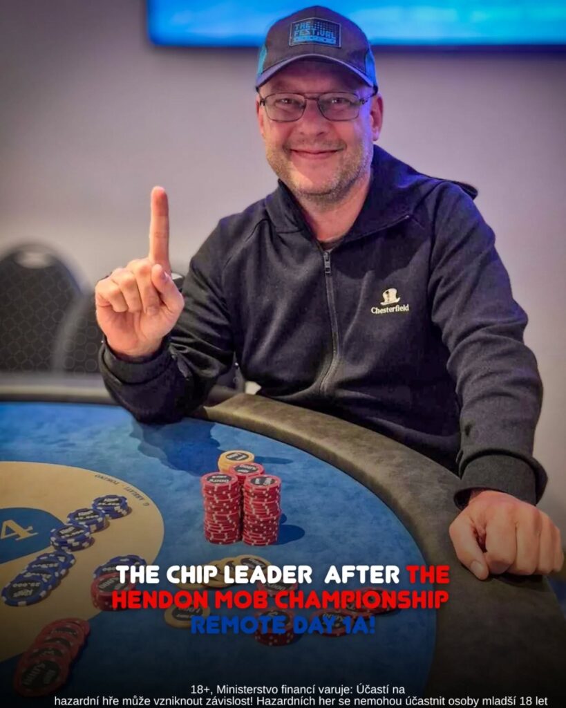 Ein Mann mit dunklen Haaren und Bart sitzt an einem Pokertisch und hält zwei Karten in der Hand. Er trägt ein schwarzes Hemd und eine schwarze Kappe. Im Hintergrund sind Pokerchips und