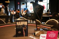 Ein Mann und eine Frau sitzen lächelnd an einem Pokertisch, während sie Karten halten. Die Frau trägt eine schwarze Brille und der Mann hat einen Bart. Im Hintergrund sind Pokerchips und Getr