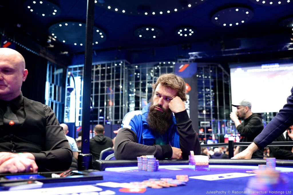 Ein Mann spielt Poker mit einem ernsten Gesichtsausdruck, während er Karten auf einem Tisch hält. Im Hintergrund sind andere Spieler und Pokerchips zu sehen.