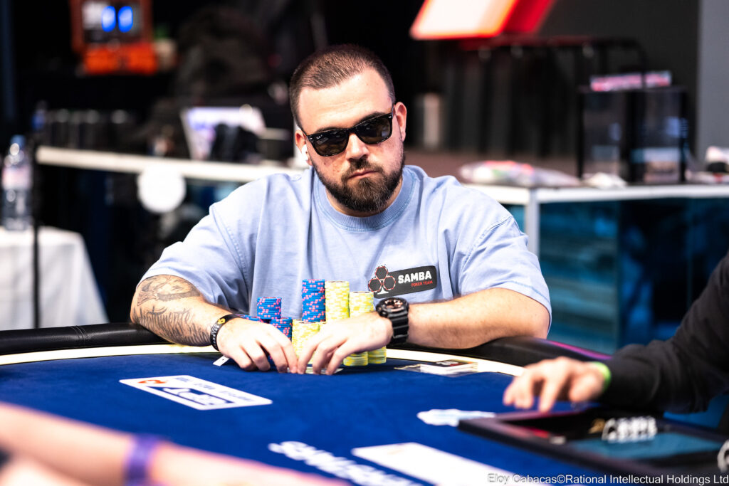 Ein Mann sitzt an einem Pokertisch und hält zwei Spielkarten in der Hand. Er schaut konzentriert auf den Tisch, umgeben von anderen Spielern. Ein Stapel Pokerchips liegt vor ihm.