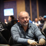 Ein Mann und eine Frau sitzen an einem Pokertisch in einem luxuriösen Casino. Der Mann hat Karten in der Hand und beide haben konzentrierte Gesichtsausdrücke. Im Hintergrund sind andere Spieler