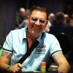 Ein Mann sitzt an einem Pokertisch und hält zwei Spielkarten in der Hand. Er hat einen konzentrierten Blick und trägt ein dunkles Hemd. Im Hintergrund sind weitere Spieler und Pokerchips zu sehen