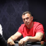 Ein Mann sitzt an einem Pokertisch mit grüner Filzdecke und hält zwei Spielkarten in der Hand. Er hat konzentrierten Blick auf die Karten und trägt eine schwarze Kappe sowie ein dunk