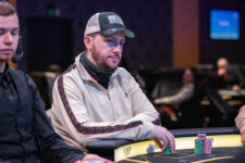Ein Mann mittleren Alters mit dunklen Haaren und einem gestutzten Bart sitzt an einem Pokertisch und hält Karten in seiner Hand. Er trägt ein dunkles Hemd und konzentriert sich auf das