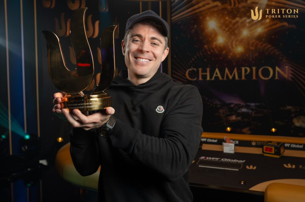 Ein Mann in einem schwarzen Anzug und einer schwarzen Sonnenbrille, umgeben von anderen Pokerspielern an einem Tisch. Er hält eine große Trophäe in die Luft und lächelt siegreich.