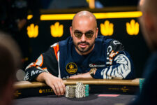 Ein Mann sitzt an einem Pokertisch während eines Turniers. Er hat einen konzentrierten Ausdruck und trägt eine schwarze Kappe. Im Hintergrund sind andere Spieler und Zuschauer zu sehen.