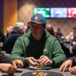 Ein Spieler sitzt an einem Pokertisch, hält Karten in der Hand und schaut konzentriert auf den Tisch. Um ihn herum sind Pokerchips und andere Spieler zu sehen.