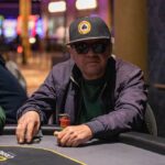Ein Mann und eine Frau spielen Poker an einem eleganten Tisch mit grüner Filzdecke. Der Mann hat eine Hand mit Karten in der Luft und schaut konzentriert auf sein Blatt, während die Frau mit ges