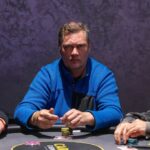 Ein Mann in einem schwarzen Anzug sitzt an einem Pokertisch und hält zwei Spielkarten in der Hand, während er konzentriert auf seine Gegner blickt.