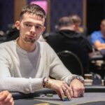 Ein Mann sitzt an einem Pokertisch und hält zwei Spielkarten in der Hand. Er trägt eine schwarze Kappe und hat eine ernste Miene. Im Hintergrund sind weitere Spieler und Pokerchips zu sehen.