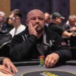 Ein Mann in einem eleganten schwarzen Anzug und einer roten Krawatte sitzt an einem Pokertisch mit grünem Filz. Er hält zwei Spielkarten in der Hand und schaut konzentriert auf die