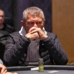 Ein Mann sitzt an einem Pokertisch und hält zwei Spielkarten in der Hand. Gegenüber von ihm sitzt eine Frau, die ebenfalls Karten hält. Im Hintergrund sind Pokerchips und Getränke