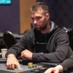 Ein professioneller Pokertisch mit grünem Filz und einem Stapel von Pokerchips in verschiedenen Farben, umgeben von Spielkarten und Getränken. Im Hintergrund sind unscharfe Personen zu sehen, die