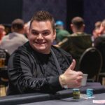 Ein Mann in einem dunkelblauen Anzug sitzt an einem Pokertisch und hält zwei Spielkarten in der Hand. Er schaut konzentriert auf seine Karten, während im Hintergrund andere Spieler und Pokerch