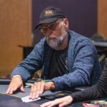 Ein Mann sitzt an einem Pokertisch und hält seine Karten in der Hand, während er konzentriert auf den Tisch schaut. Im Hintergrund sind weitere Spieler und Pokerchips zu sehen.