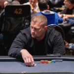 Ein Mann und eine Frau spielen Poker an einem eleganten Tisch in einem stilvollen Raum. Der Mann hält Karten in der Hand, während die Frau konzentriert auf ihre Karten schaut. In der Mitte des T