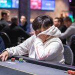Ein Mann sitzt an einem Pokertisch und hält zwei Spielkarten in der Hand. Er trägt eine dunkle Jacke und hat eine ernste Miene. Im Hintergrund sind weitere Spieler und Spielchips zu sehen.