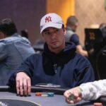 Ein Mann in einem dunkelblauen Anzug und einer roten Krawatte sitzt an einem Pokertisch mit Chips und Karten. Er hält zwei Spielkarten in der Hand und betrachtet sie konzentriert.