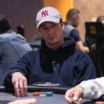 Ein Mann in dunkler Kleidung sitzt an einem Pokertisch und hält in der Hand Spielkarten. Er wirkt konzentriert und fokussiert. Im Hintergrund sind weitere Spieler und Pokerchips zu