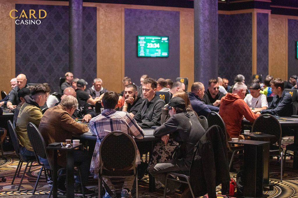 Ein Mann in einem dunkelblauen Anzug und weißem Hemd sitzt an einem Pokertisch und hält eine Hand mit Spielkarten. In der Mitte des Tisches liegen weitere Karten und Jetons. Im H