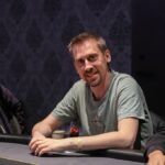 Ein Mann sitzt an einem Pokertisch und hält zwei Spielkarten in der Hand. Er trägt eine schwarze Kappe und eine dunkle Sonnenbrille. Auf dem Tisch liegen Pokerchips und eine Flas