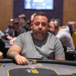 Ein Mann mit schwarzer Sonnenbrille und einem roten Hemd sitzt an einem Pokertisch und hält zwei Spielkarten in der Hand. Er wirkt konzentriert und fokussiert auf das Spiel. Im