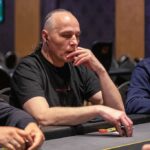 Ein Mann sitzt an einem Pokertisch, umgeben von anderen Spielern. Er hält seine Karten in der Hand und schaut konzentriert auf den Tisch. Die anderen Spieler haben ebenfalls Karten in der Hand und Chips