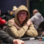 Ein Mann mit schwarzen Haaren und Bart sitzt lächelnd an einem Pokertisch und hält eine Hand mit Spielkarten. Er trägt ein dunkles Hemd und eine Brille. Im Hintergrund sind weitere Spieler