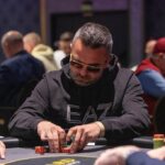 Ein professioneller Pokertisch mit grünem Filz, umgeben von Stühlen und Chips. Zwei Personen spielen Karten, während andere zuschauen. Die Atmosphäre ist gespannt und konzentriert.
