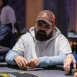 Ein Mann sitzt an einem Pokertisch und hält zwei Spielkarten in der Hand. Er schaut konzentriert auf den Tisch, umgeben von Pokerchips und anderen Spielern.