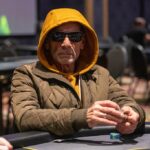 Ein Mann sitzt an einem Pokertisch und hält zwei Spielkarten in der Hand. Er wirkt konzentriert und umgeben von anderen Pokerspielern.