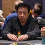 Ein Mann sitzt an einem Pokertisch und betrachtet konzentriert seine Karten. Im Hintergrund sind andere Spieler und Spielchips zu sehen.