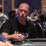 Ein Mann in einem dunkelgrauen Anzug sitzt an einem Pokertisch und hält seine Karten in der Hand, während er konzentriert auf seine Gegner schaut. Die Spannung des Pokerspiels ist in