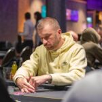 Ein Mann sitzt an einem Pokertisch und hält zwei Spielkarten in der Hand, während er konzentriert auf seine Gegner schaut. Die Chips auf dem Tisch sind bunt und stapeln sich hoch.