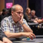 Ein professioneller Pokerspieler mit Sonnenbrille und Kappe betrachtet konzentriert seine Karten während eines Turniers.