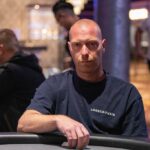 Ein Mann und eine Frau spielen Poker an einem Tisch, sie halten Karten in der Hand und schauen konzentriert auf ihre Chips. Die Frau hat blondes Haar und trägt ein rotes Oberteil, der Mann hat