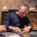 Ein Mann sitzt an einem Pokertisch und hält zwei Karten in der Hand. Er schaut konzentriert auf den Tisch, auf dem Pokerchips und Karten liegen. In der Hintergrund sieht man weitere