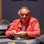 Ein Mann spielt Poker an einem Tisch mit grünem Filz, umgeben von anderen Spielern. Er hält Karten in der Hand und schaut konzentriert auf den Tisch.