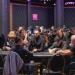 Ein Mann spielt Poker an einem Tisch in einem eleganten Casino. Sein Gesichtsausdruck ist konzentriert, während er seine Karten betrachtet. Im Hintergrund sind andere Spieler und Casinomitar