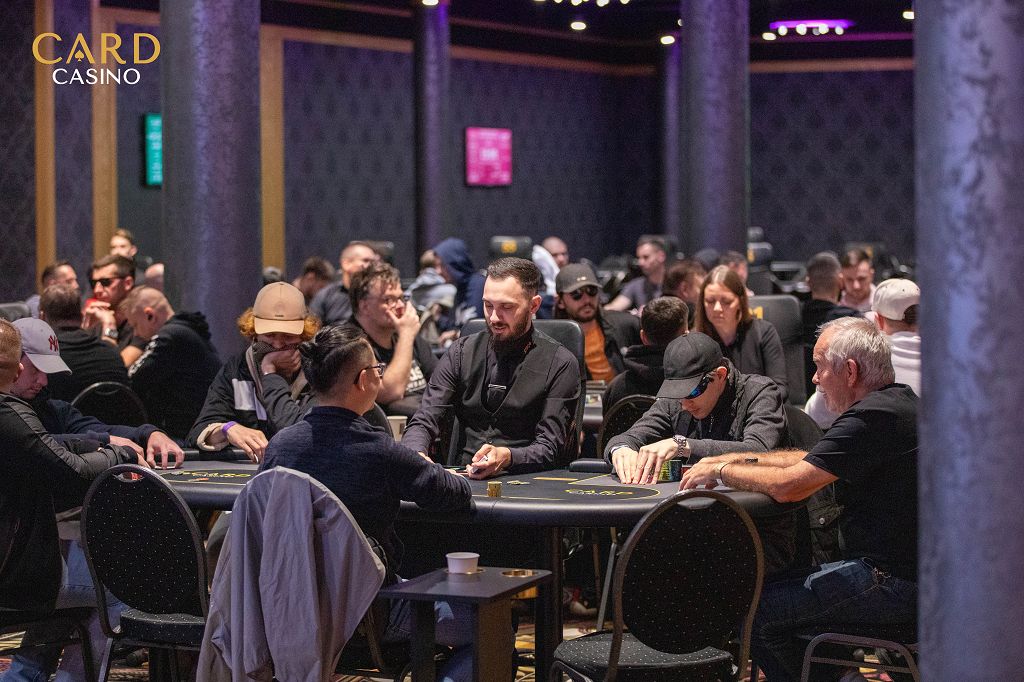 Ein Mann spielt Poker an einem Tisch in einem eleganten Casino. Sein Gesichtsausdruck ist konzentriert, während er seine Karten betrachtet. Im Hintergrund sind andere Spieler und Casinomitar